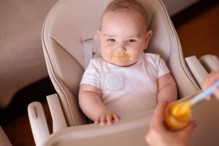 Photo for High angle view of mother feeding cheerful baby boy with porridge, baby sitting in high chair all messy and staied smiling and eating - Royalty Free Image