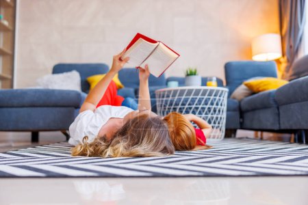 Téléchargez les photos : Mère et fille couchées sur le sol se relaxant à la maison ensemble, mère lisant un livre à sa petite fille - en image libre de droit