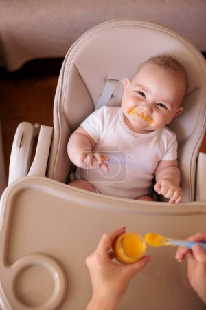 Foto de Vista de ángulo alto de la madre alimentando al niño alegre con gachas, el bebé sentado en la silla alta todo desordenado y estable sonriendo y comiendo - Imagen libre de derechos