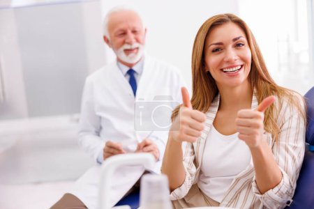 Photo for Portrait of satisfied patient sitting at dental chair at dentist office, smiling and showing thumbs up with doctor in the background - Royalty Free Image
