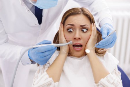 Téléchargez les photos : Femme effrayée alors qu'elle est assise à la chaise dentaire au cabinet du dentiste pendant que le médecin tient une perceuse dentaire et un miroir incliné, réparant la dent du patient - en image libre de droit