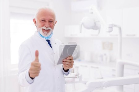 Téléchargez les photos : Portrait d'un médecin principal portant l'uniforme debout à l'hôpital montrant les pouces levés lors de la vérification des dossiers médicaux à l'aide d'un ordinateur tablette - en image libre de droit
