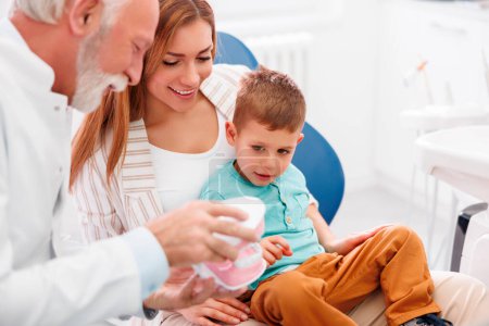 Photo for Child having a regular checkup at dentist office, sitting at mother's lap while senior male dentist is demonstrating procedure on plastic jaw model - Royalty Free Image