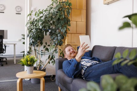 Téléchargez les photos : Jeune homme d'affaires occasionnel allongé sur le canapé au bureau, ayant un appel vidéo à l'aide d'un ordinateur tablette - en image libre de droit