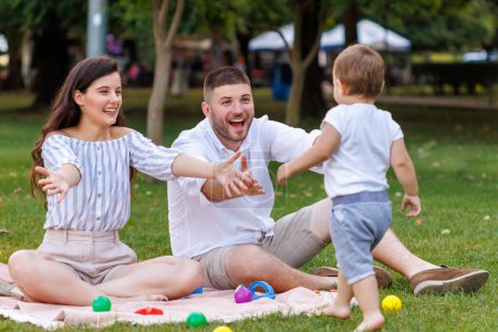 Foto de Niño lindo juguetón corriendo hacia sus padres mientras hacen picnic en el parque; madre y padre sentados en una manta de picnic divirtiéndose jugando con su hijo - Imagen libre de derechos