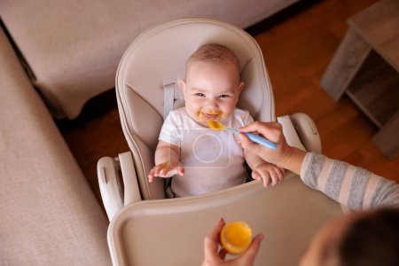 Vue grand angle de la mère nourrissant gai bébé garçon avec de la bouillie, bébé assis dans la chaise haute tout désordonné et staied souriant et mangeant