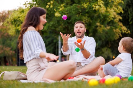 Téléchargez les photos : Beaux jeunes parents pique-niquer dans le parc avec leur mignon petit garçon, assis sur la couverture et jouant avec des boules colorées - en image libre de droit