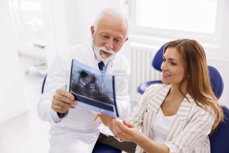 Téléchargez les photos : Dentiste expliquant les procédures nécessaires au patient assis dans une chaise dentaire tout en regardant la radiographie de la mâchoire à la clinique de stomatologie - en image libre de droit