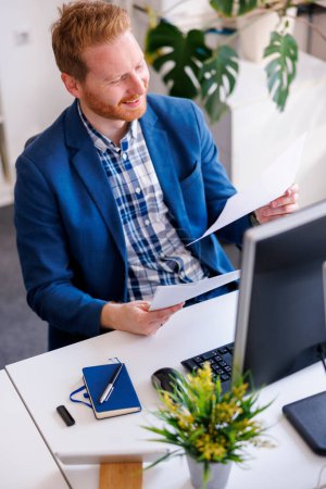 Photo for High angle view of young successful businessman sitting at his desk analysing financial reports while working in an office - Royalty Free Image