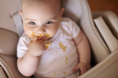 Téléchargez les photos : Vue grand angle de mignon petit garçon sucer doigt après avoir mangé du porridge dans la chaise haute, regardant vers la caméra et souriant - en image libre de droit
