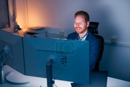 Photo for Confident successful businessman sitting at his desk using desktop computer while working late in an office, smiling and looking towards computer screen - Royalty Free Image