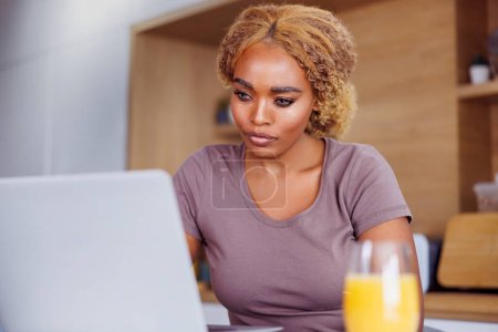 Téléchargez les photos : Femme freelance assis au comptoir de cuisine prendre le petit déjeuner et de travailler à la maison à l'aide d'un ordinateur portable - en image libre de droit