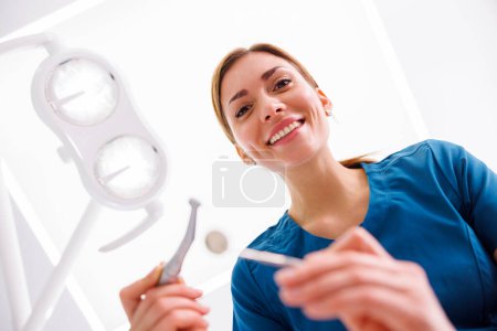Photo for Low angle view of female dentist holding dental drill and angled mirror while fixing patients tooth - Royalty Free Image