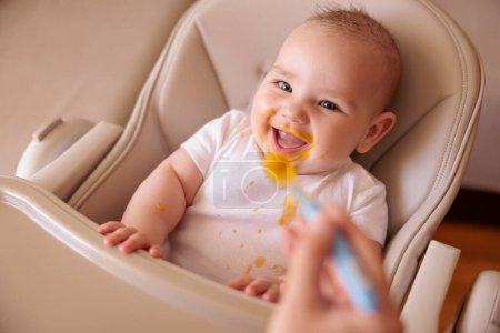 High angle view of mother feeding cheerful baby boy with porridge using spoon, baby sitting in high chair all messy and staied smiling and eating