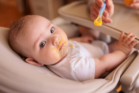 Mère nourrissant son bébé garçon à l'aide d'une cuillère, introduisant le premier repas solide, bébé assis dans une chaise haute tout salissant et empilé de bouillie, regardant vers la caméra