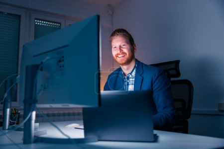 Foto de Hombre de negocios exitoso y seguro sentado en su escritorio usando una computadora de escritorio mientras trabaja hasta tarde en una oficina, sonriendo y mirando hacia la pantalla de la computadora - Imagen libre de derechos