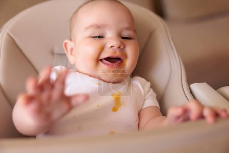 Foto de Lindo bebé niño riendo y jugando en la silla alta todo desordenado y manchado después de comer gachas - Imagen libre de derechos