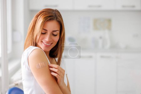 Téléchargez les photos : Portrait d'une jeune femme assise dans un établissement de santé avec un patch médical sur le bras après la vaccination - concept de vaccination, d'immunisation et de prévention des maladies - en image libre de droit