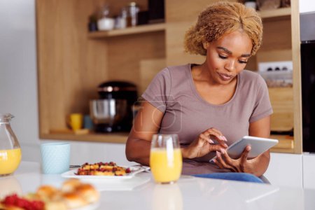 Téléchargez les photos : Jeune femme assise au comptoir de la cuisine, utilisant un ordinateur tablette tout en prenant le petit déjeuner à la maison le matin - en image libre de droit