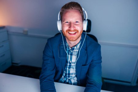 Foto de Hombre de negocios sentado en su escritorio con auriculares que tienen reunión en línea mientras trabaja hasta tarde en una oficina, sonriendo y saludando a la cámara - Imagen libre de derechos