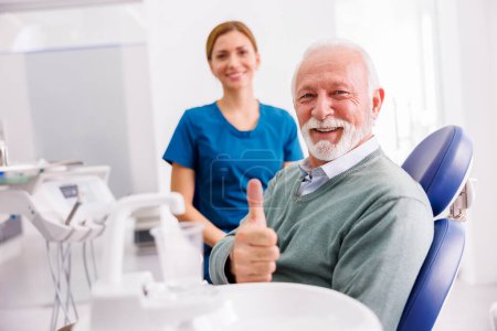 Foto de Retrato de un paciente sénior satisfecho sentado en la silla dental en el consultorio del dentista mostrando los pulgares hacia arriba y sonriendo con el médico en el fondo - Imagen libre de derechos