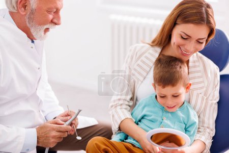 Foto de Odontólogo masculino mayor fijando el diente a un niño valiente sentado en la silla del dentista en el regazo de la madre usando taladro dental y espejo en ángulo - Imagen libre de derechos