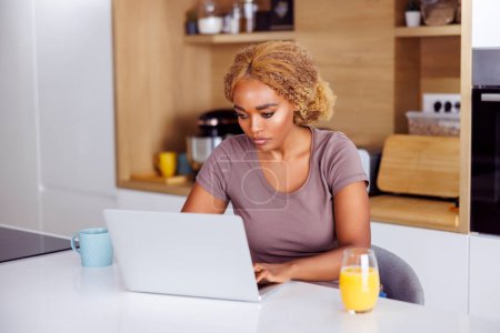 Photo for Woman sitting at kitchen counter having breakfast and working remotely from home using laptop computer - Royalty Free Image