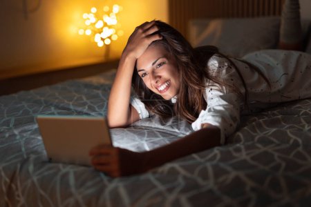 Foto de Hermosa mujer joven acostada en la cama tarde en la noche, relajándose y divirtiéndose usando tableta - Imagen libre de derechos