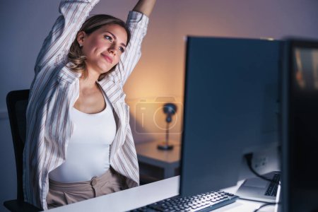 Téléchargez les photos : Femme s'étirant tandis qu'elle était assise à son bureau, fatiguée en faisant des heures supplémentaires tard dans la nuit - en image libre de droit