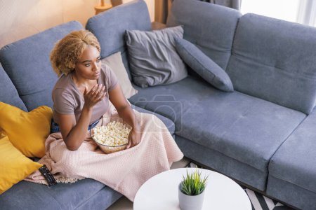 Téléchargez les photos : Vue grand angle de belle jeune femme africaine assise sur le canapé du salon, mangeant du pop-corn et regardant un film d'horreur à la télévision, choquée et effrayée - en image libre de droit