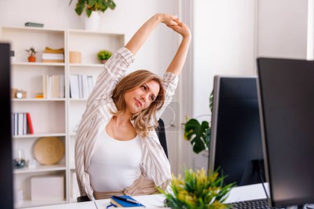 Photo for Woman stretching out while sitting at her office desk, tired after a long day at work - Royalty Free Image