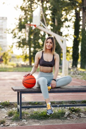 Foto de Fit young woman in sportswear sitting on a bench by an outdoor basketball court, holding a ball and relaxing while taking a workout break - Imagen libre de derechos