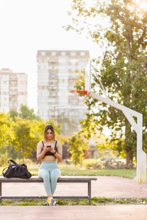 Téléchargez les photos : Jeune femme active en vêtements de sport assis sur le banc près du terrain de basket surfer sur le net en utilisant un téléphone intelligent tout en prenant une pause d'entraînement - en image libre de droit