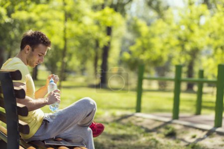 Young athlete, sitting on a wooden bench in a park next to an outdoor gym, drinking water after hard workout