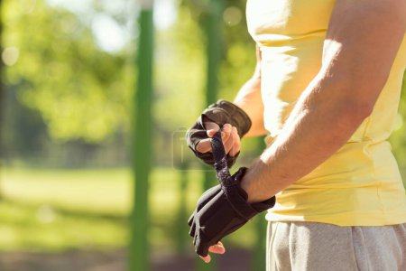 Téléchargez les photos : Muscular built young athlete working out in an outdoor gym, puts on sports gloves before training - en image libre de droit