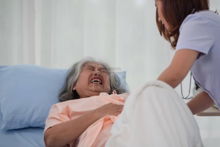 Photo for Asian nurse helping a senior Asian female patient with stomach ache on a bed in hospital ward with diagnosis or treatment, treating elderly woman - Royalty Free Image