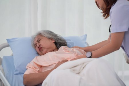 Foto de Enfermera asiática ayudando a una paciente asiática mayor con dolor de estómago en una cama en la sala de hospital con diagnóstico o tratamiento, tratando a una mujer mayor - Imagen libre de derechos