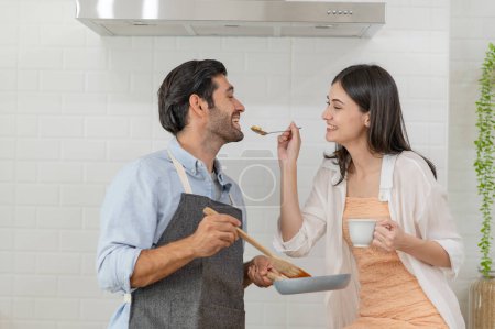 Foto de Feliz pareja joven cocinando juntos en el mostrador de la cocina, alimentándose mutuamente en su cocina en casa - Imagen libre de derechos