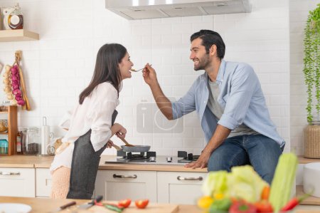Foto de Feliz pareja joven cocinando juntos en el mostrador de la cocina, alimentándose mutuamente en su cocina en casa - Imagen libre de derechos
