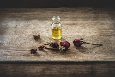 Ätherisches Öl in einer kleinen Glasflasche mit getrockneten rosa Rosen in der Knospe auf einem Holzgrund. Selektiver Fokus und Kopierraum für Text. Naturkosmetik.