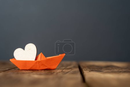 close-up of an orange miniature origami ship with a wooden heart on wooden table. dark background. abstract concept