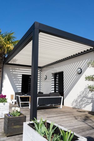 Téléchargez les photos : Eco friendly structure d'ombre pergola en aluminium bioclimatique, auvent et toit patio, salon de jardin, chaises, barbecue en métal entouré d'aménagement paysager - en image libre de droit