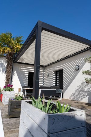 Téléchargez les photos : Eco friendly structure d'ombre pergola en aluminium bioclimatique, auvent et toit patio, salon de jardin, chaises, barbecue en métal entouré d'aménagement paysager - en image libre de droit