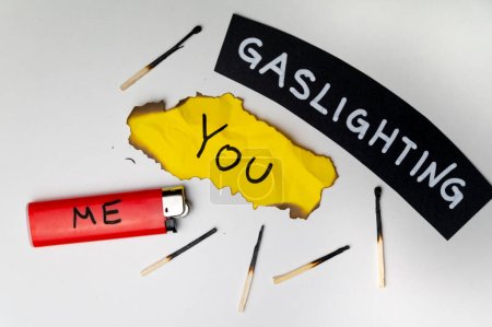 Word Gaslighting, auf einer schwarzen Fläche, neben einem Feuerzeug mit dem Wort Ich und einer verbrannten gelben Karte mit dem Schriftzug Du. Psychologische Bedeutung.