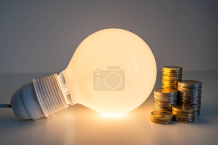 Light bulb turned on, with stacks of coins next to it. Rising electricity tariffs, energy dependency, energy sources and energy supplies.