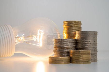 Téléchargez les photos : Light bulb turned on, with stacks of coins next to it. Rising electricity tariffs, energy dependency, energy sources and energy supplies. - en image libre de droit