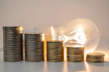 Foto de Lámpara de luz con pilas de monedas de altura descendente. Reducción de las tarifas energéticas. - Imagen libre de derechos