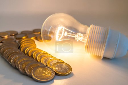 Glühbirne angezündet, daneben eine Reihe Münzen. Trends bei Strompreisen, Energieabhängigkeit, Energieversorgung.