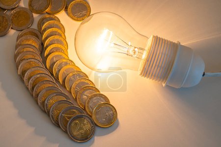Glühbirne angezündet, daneben eine Reihe Münzen. Trends bei Strompreisen, Energieabhängigkeit, Energieversorgung.