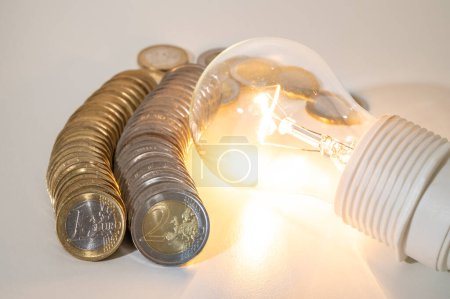 Photo for Light bulb lit, with row of coins next to it. Trends in electricity tariffs, energy dependence, energy supplies. - Royalty Free Image